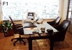 F1 – Aqui vê-se uma mesa com três cadeiras escuras, uma delas com espaldar mais alto, braços, rodas e elementos de regulagem. Sobre a mesa, objetos como monitor de tela plana, telefone e apoio de couro para escrever. Ao fundo, uma grande janela
