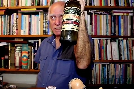 Ignácio de Loyola Brandão em seu escritório segurando garrafa da aguardente Providência, caprichosamente engarrafada no passado pelos Irmãos Guerra de Araraquara