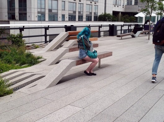 A moça de cabelo azul senta-se no banco público e observa com cuidado a sacola de papel; o detetive novato tenta fotografar seu rosto, mas sem sucesso.