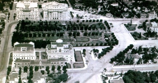 Foto aérea da Praça da Liberdade em 1934, mostrando a Alameda Central com suas Palmeiras Imperiais, as quatro Secretarias de Estado e o Palácio da Liberdade (à direita). Destaque para o terreno triangular (acima) onde então se assentava o Palacete Dolabel