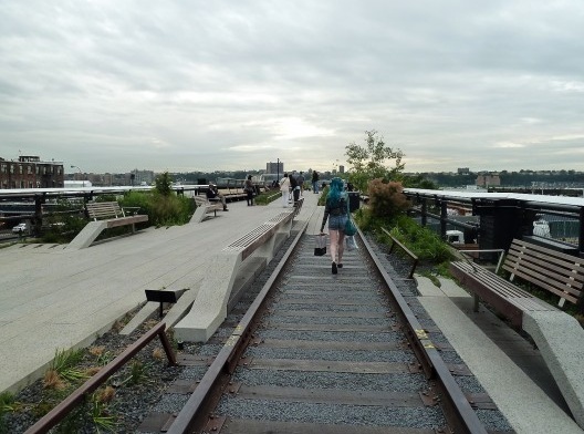 Carregando sua bolsa e uma sacola de papelão, a moça de cabelo azul caminha rápido no dia nublado.