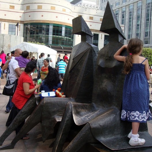 Recostada em uma estátua, uma graciosa menina observa distraída a praça movimentada.