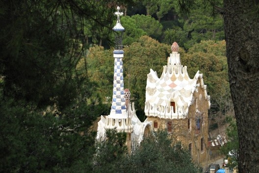 Coberturas dos pórticos da entrada principal do Park Güell, adornadas com elementos da natureza (musgo, troncos, cogumelos). Notar a pujança da vegetação ao redor, onde predominam os pinheiros.