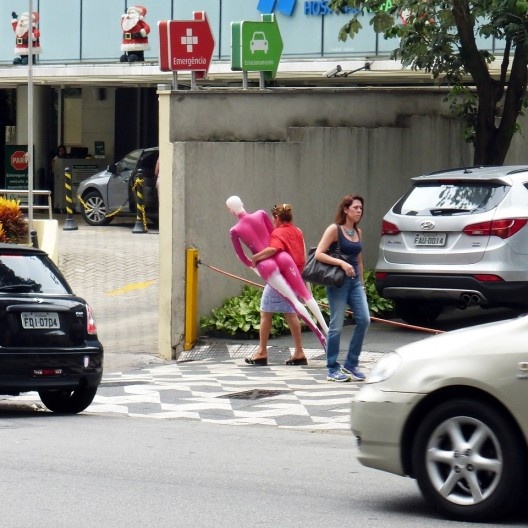 Com passos determinados, uma mulher segue anônima no meio do burburinho da metrópole levando nos braços uma manequim.