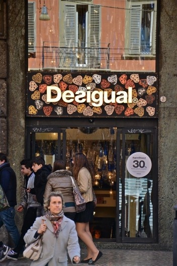 Centro Histórico de Bolonha, pedestres abrigados em pórtico e vitrine com publicidade de loja